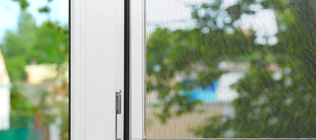 Close-up of a white window frame and fine mesh screen, with a softly blurred green garden beyond, creating a calm, airy summer feel.