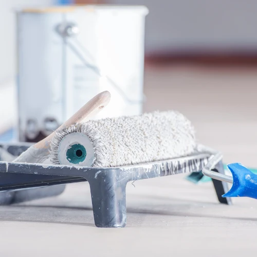 A paint roller coated in white paint rests in a metal tray, with a blue-handled tool beside it and a paint bucket blurred behind, suggesting a calm home renovation.