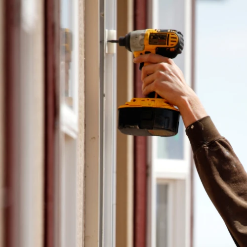 A worker uses a yellow cordless drill to secure a white window frame, with daylight streaming through the glass; the scene feels focused and practical.