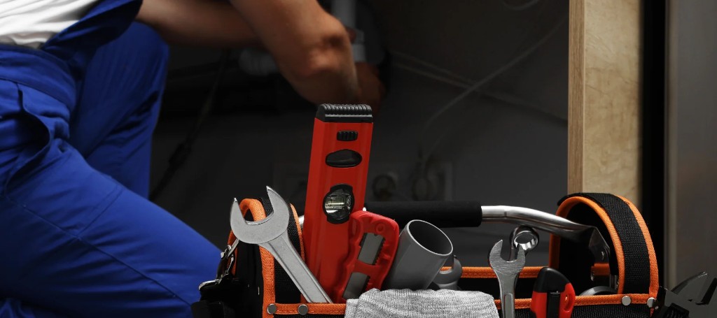 A tradesperson in blue workwear kneels beside an open black-and-orange tool bag filled with wrenches and a red power tool in a dim workshop.