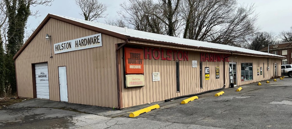 Holston Hardware, a long, tan-sided rural store with red signage, sits beside an empty, weathered parking lot under bare winter trees.