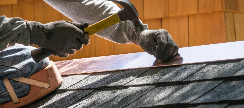 A roofer in work gloves hammers a metal strip onto dark shingles, carefully repairing a roof in warm sunlight; the scene feels focused and industrious.