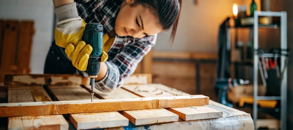A focused woodworker in yellow gloves drills into a plank on a workbench in a warmly lit workshop, creating a careful, hands-on scene.