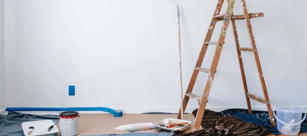 A wooden A-frame ladder, paint bucket, roller, and tools sit on a dark drop cloth in a bright white room, suggesting calm, ongoing renovation.