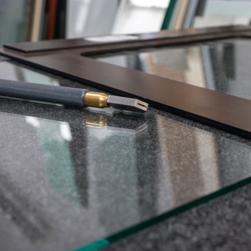 A glass cutter with a textured black handle and brass collar rests on reflective glass beside dark framing, suggesting precise window or pane cutting.