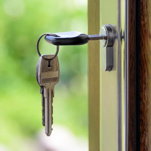 A metal key inserted in a window lock, with a second key hanging from its ring; soft green foliage outside creates a calm, secure atmosphere.